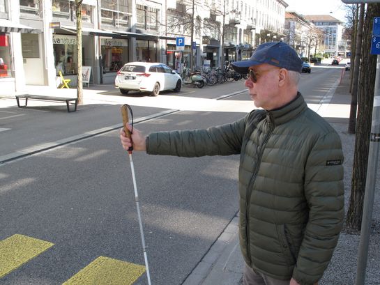 Gerd Bingemann hält seinen weissen Stock am Zebrastreifen heraus, um darauf aufnerksam zu machen, dass er die Strasse überqueren möchte. 