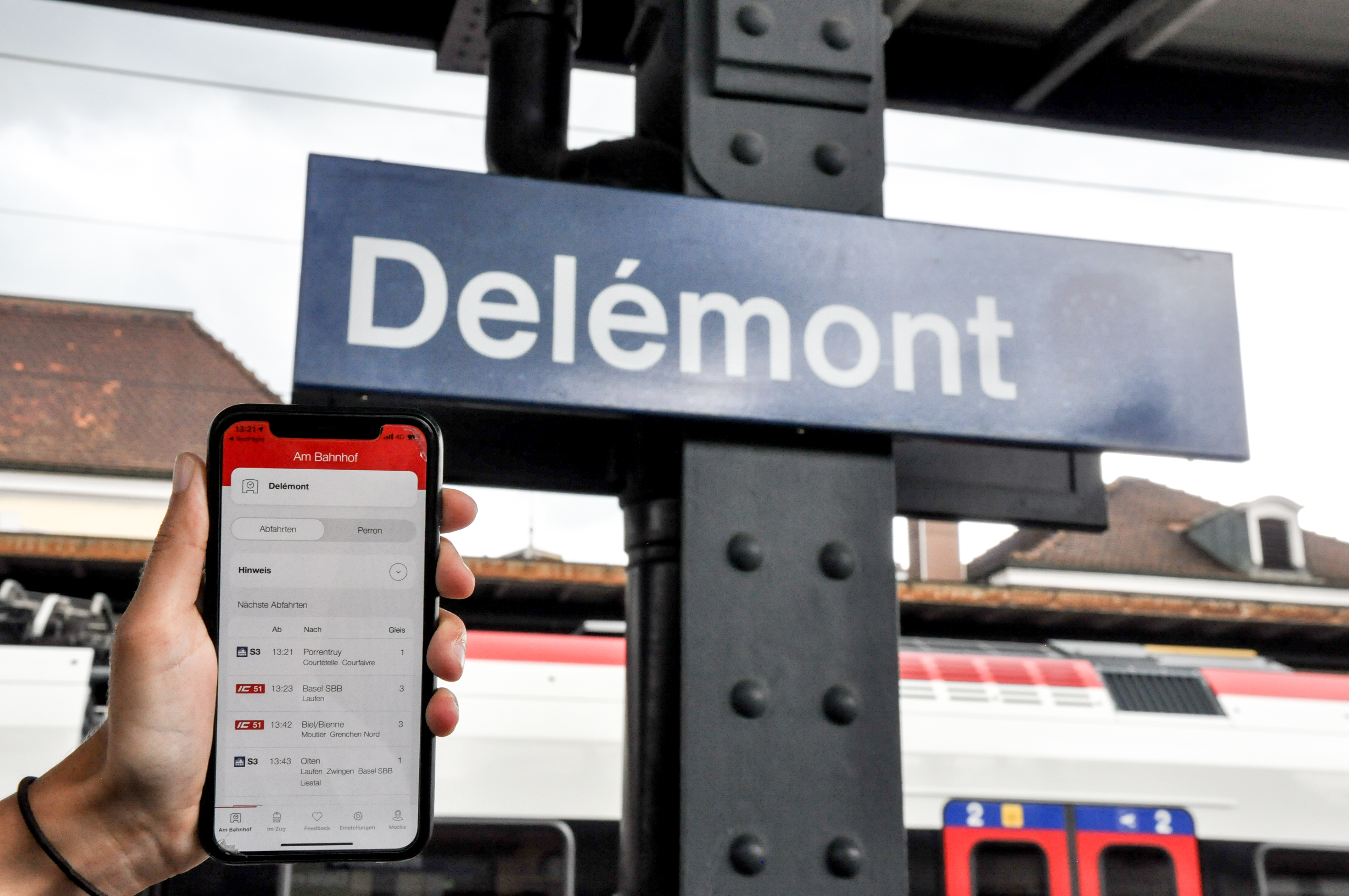 Vor einem Schild der SBB, auf dem die Stadt Delémont angegeben ist, befindet sich ein Smartphone, auf dem die Anwendung geöffnet ist.